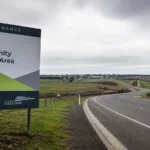 Roadside sign for Lovely Banks future community growth area beside open development land and a new road, highlighting tension between housing supply and infrastructure.