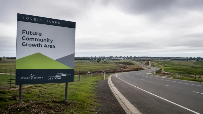 Roadside sign for Lovely Banks future community growth area beside open development land and a new road, highlighting tension between housing supply and infrastructure.