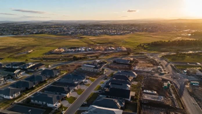 Aerial view of a large Australian housing estate expanding into greenfield land, with completed homes, new roads and houses under construction at sunset. 