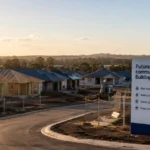 A new Australian housing estate under construction at sunset, with unfinished homes, roads, fencing and a sign highlighting future community infrastructure including roads, water connections and sewer upgrades.