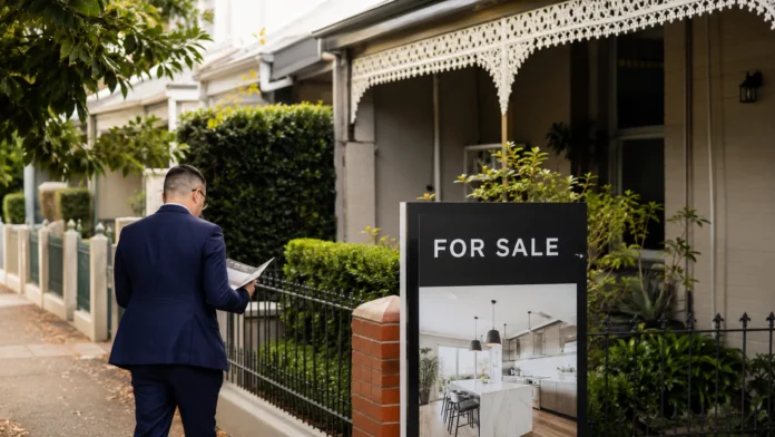 Property professional reviewing documents beside a for-sale sign outside an established Australian home, illustrating debate over housing tax changes and supply
