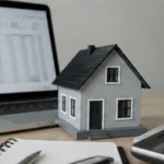 Miniature house beside a laptop showing financial charts, with a calculator and notebook on a desk.