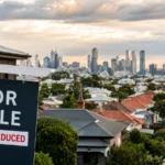 For sale sign with price reduced label overlooking Australian suburban rooftops and a city skyline, reflecting softer home prices and investor pressure.